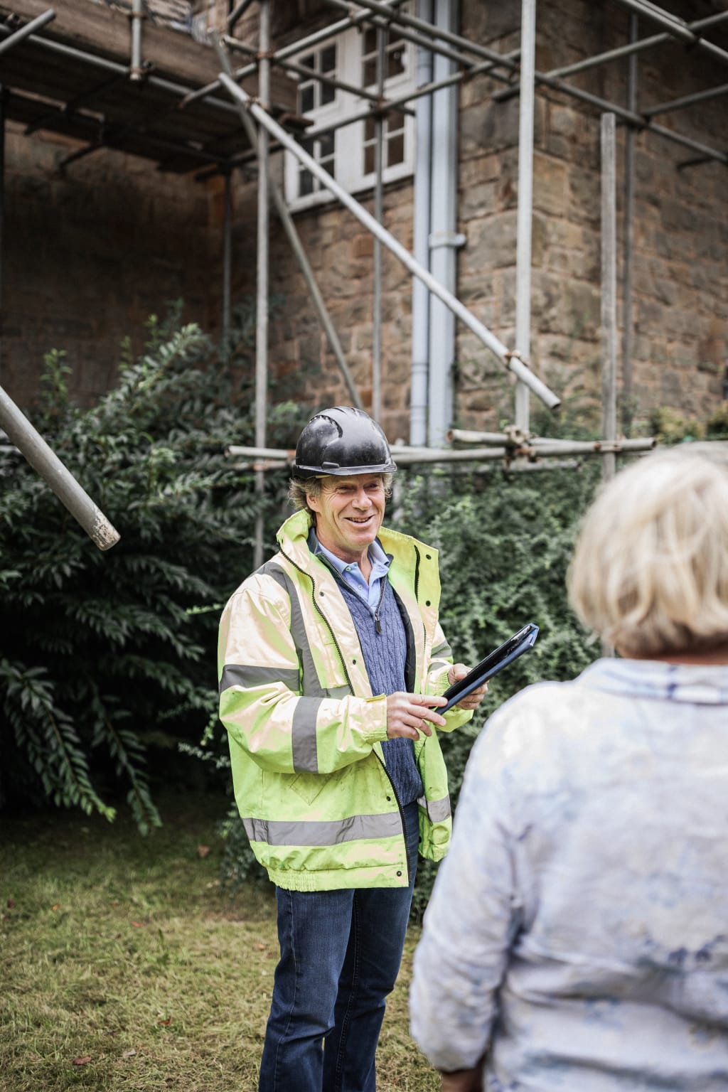 A Cymru director on a country property survey, scaffolded stonework behind, in high-vis with a tablet