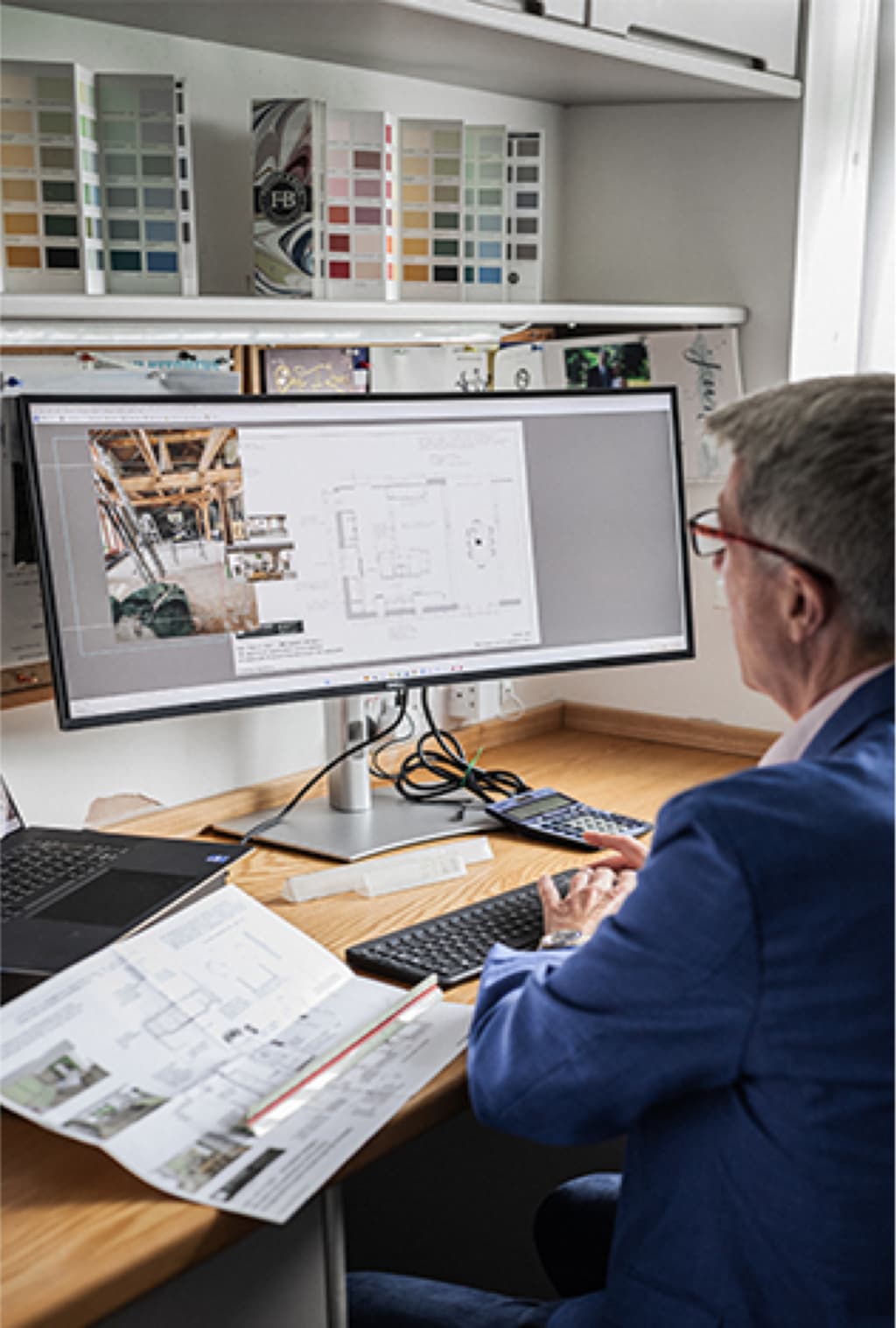 Cymru designer working on a kitchen plan at an ultrawide CAD monitor in the Newport studio, paint swatches above